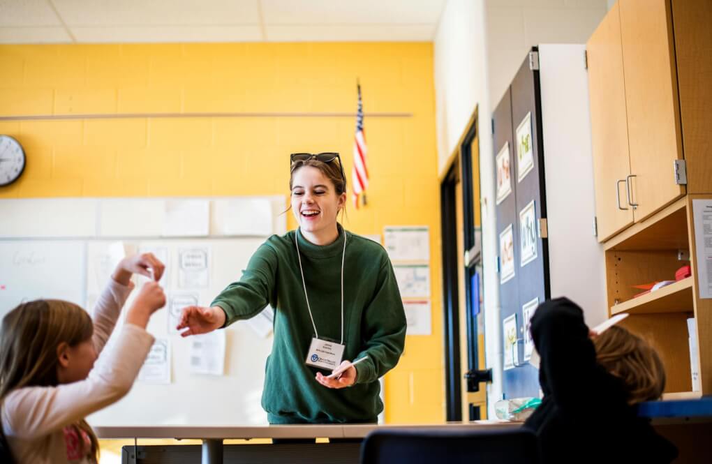 A student teacher smiles and reaches out toward and elementary school student. There is a white board in the background. Another student is blurred out in the foreground.
